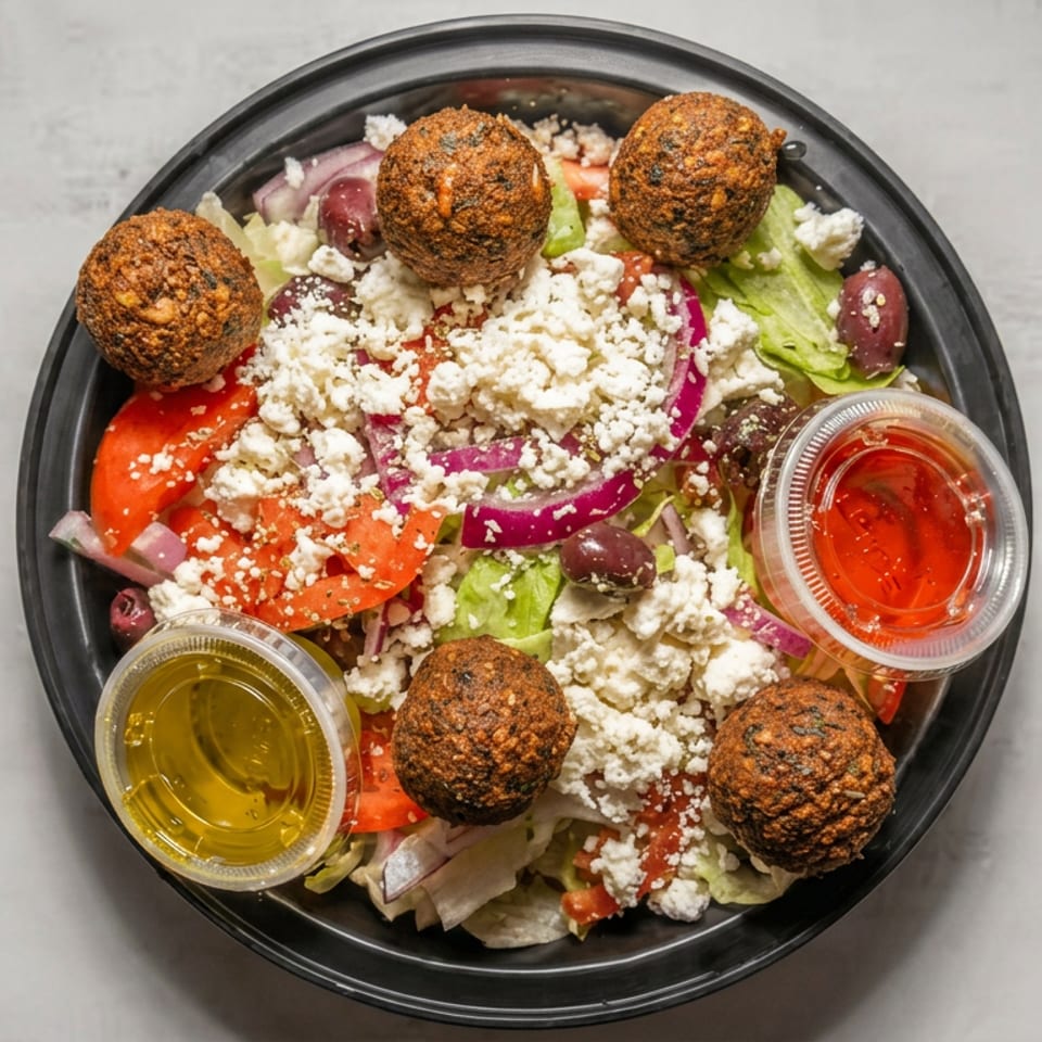Greek salad topped with five falafel balls, crumbled feta, tomatoes, onions, olives, lettuce, and cups of dressing.