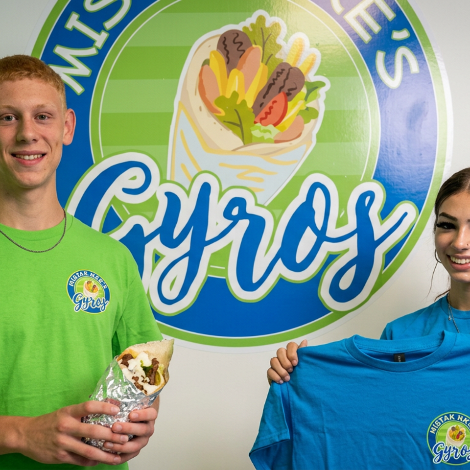 Two smiling people display a wrapped gyro and branded blue and green shirts beneath a colorful gyros restaurant logo.