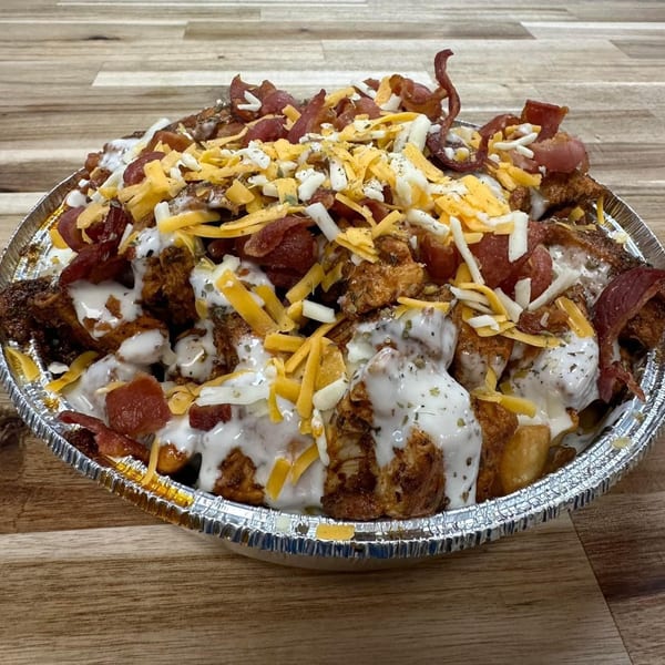Loaded fries with chicken, bacon, shredded cheese, and white sauce in a foil container.